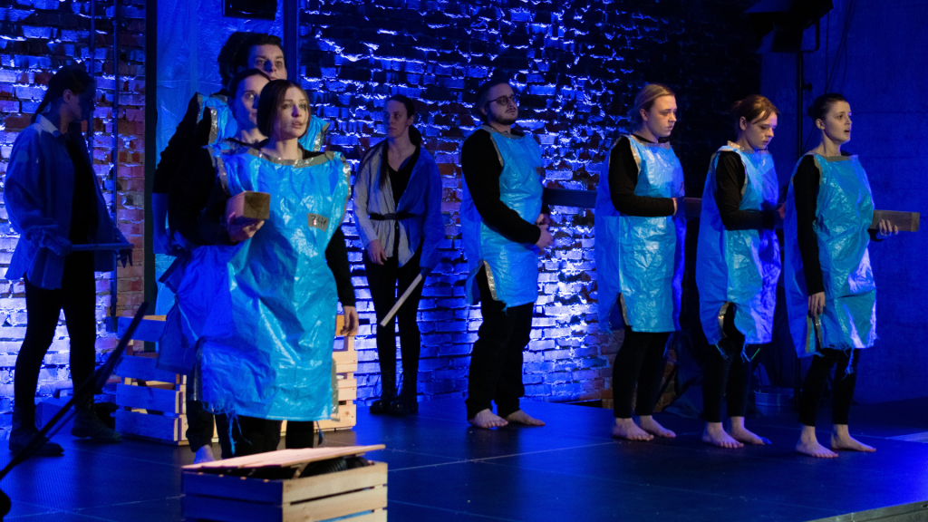 Grupa młodych aktorów na scenie podczas spektaklu, ubranych w prowizoryczne kostiumy wykonane z niebieskiej folii. Niektórzy trzymają drewniane klocki i elementy. Tło stanowi ceglana ściana podświetlona na niebiesko.