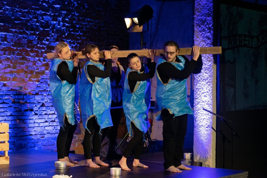 Grupa pięciu młodych aktorów ubranych w niebieskie worki imitujące więzienne uniformy niesie wspólnie drewnianą belkę na ramionach, stojąc boso na scenie. W tle ceglany mur i napis „Arbeit macht frei” widoczny w ciemnościach.