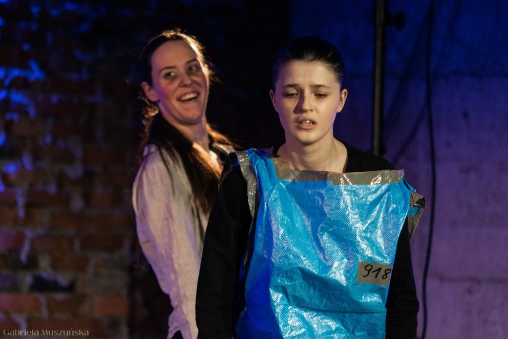 Zbliżenie na dwie aktorki – jedna z nich, w niebieskim stroju z numerem 918, wygląda na wyczerpaną i zrozpaczoną; druga, stojąca za nią, uśmiecha się ironicznie, co tworzy kontrast emocjonalny między postaciami.