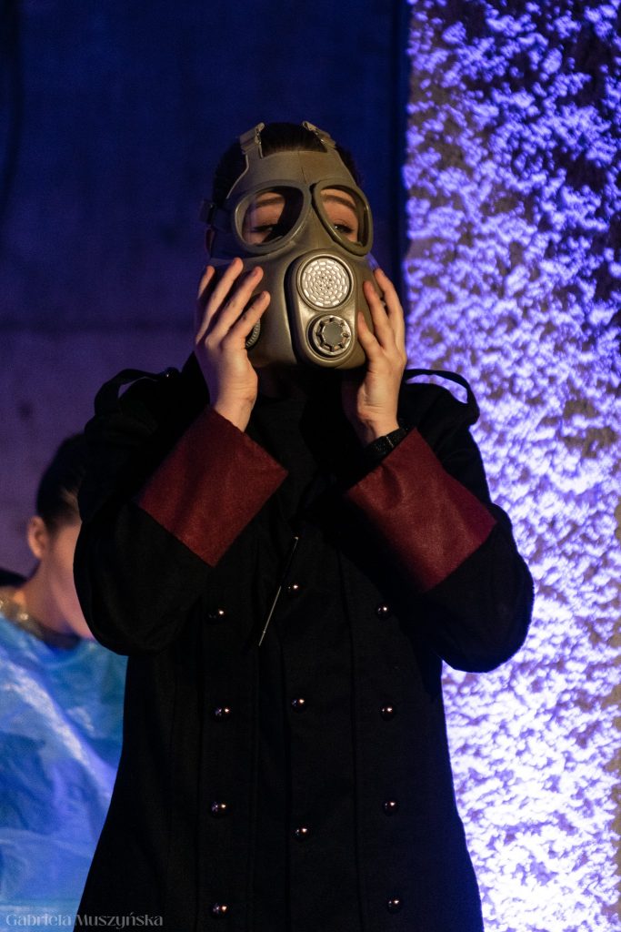 Postać ubrana w czarny płaszcz z bordowymi mankietami zakłada na twarz starą maskę gazową. W tle widoczna inna postać w niebieskim stroju, scena oświetlona jest na niebiesko-fioletowo.