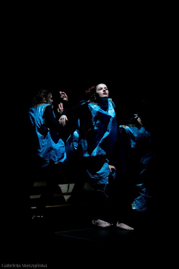 Grupa aktorek w niebieskich uniformach z folii znajduje się w półmroku. Na pierwszym planie postać z uniesioną ręką i intensywnym spojrzeniem ku górze, oświetlona dramatycznym światłem. Pozostałe sylwetki są ledwo widoczne w cieniu.