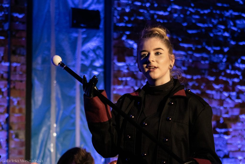 Młoda aktorka w czarnym płaszczu z bordowymi mankietami unosi czarną pałkę zakończoną białą kulką. Jej twarz wyraża pewność siebie, a scena jest oświetlona niebieskim światłem na tle ceglanej ściany.