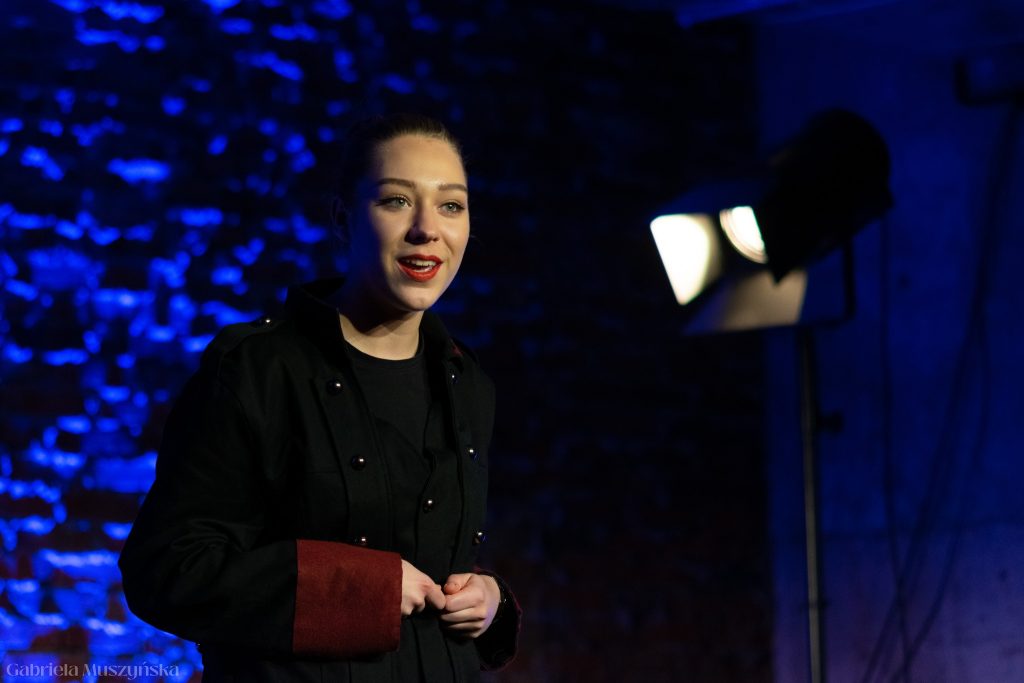 Zbliżenie na młodą aktorkę w czarnym płaszczu z bordowymi mankietami, patrzącą przed siebie z lekkim uśmiechem. W tle reflektor i ceglana ściana w niebieskim świetle.