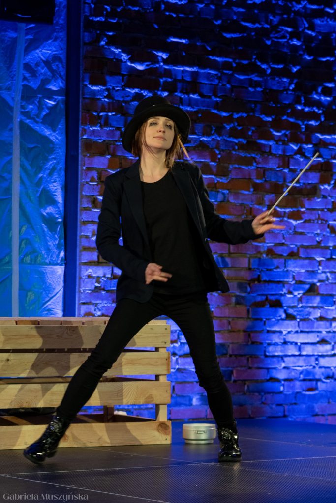 Aktorka w czarnym kapeluszu i garniturze pozuje z teatralnym rozmachem, trzymając w ręce batonik lub wskaźnik. Scena podświetlona jest niebiesko-fioletowym światłem, a za nią znajduje się drewniana skrzynia.