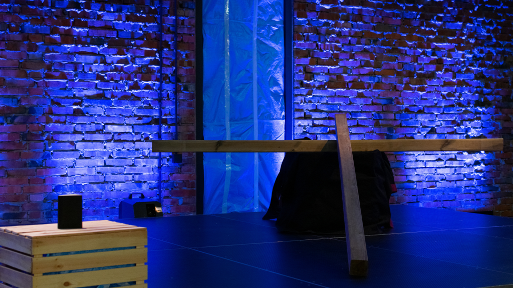 Minimalistyczna scenografia na scenie teatralnej: drewniany krzyż oparty o czarny materiał, ustawiony na tle ceglanej ściany oświetlonej niebieskim światłem. Po lewej stronie widoczna drewniana skrzynka i niewielki głośnik.
