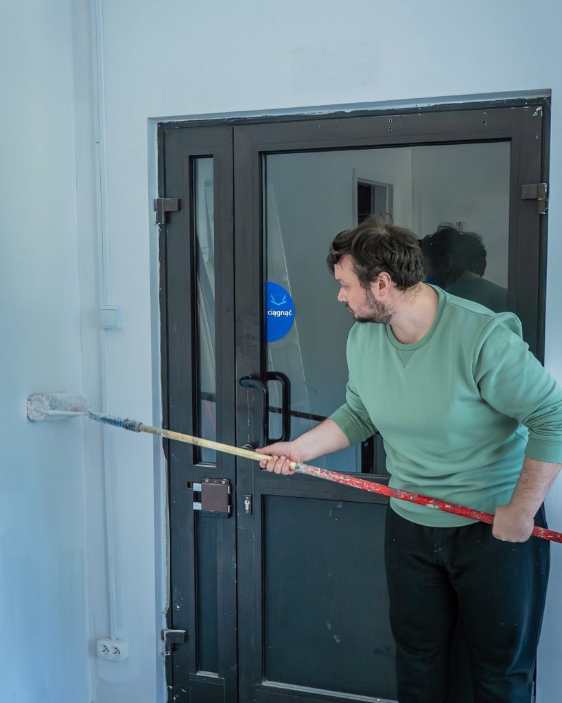 Mężczyzna w zielonej bluzie maluje biały mur za pomocą wałka na długim kiju, stojąc przy czarnych drzwiach wejściowych. Remont trwa.