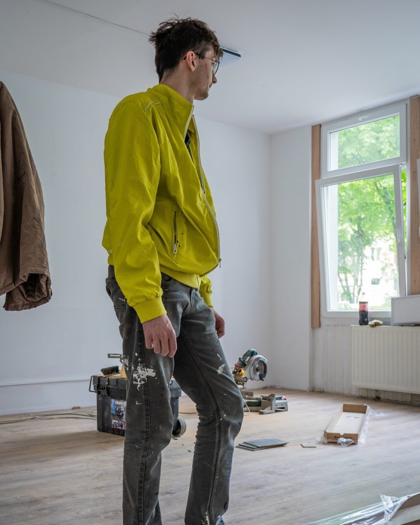 Mężczyzna w żółtej kurtce i poplamionych farbą spodniach stoi na środku remontowanego pomieszczenia, obserwując prace wykończeniowe. Remont trwa.