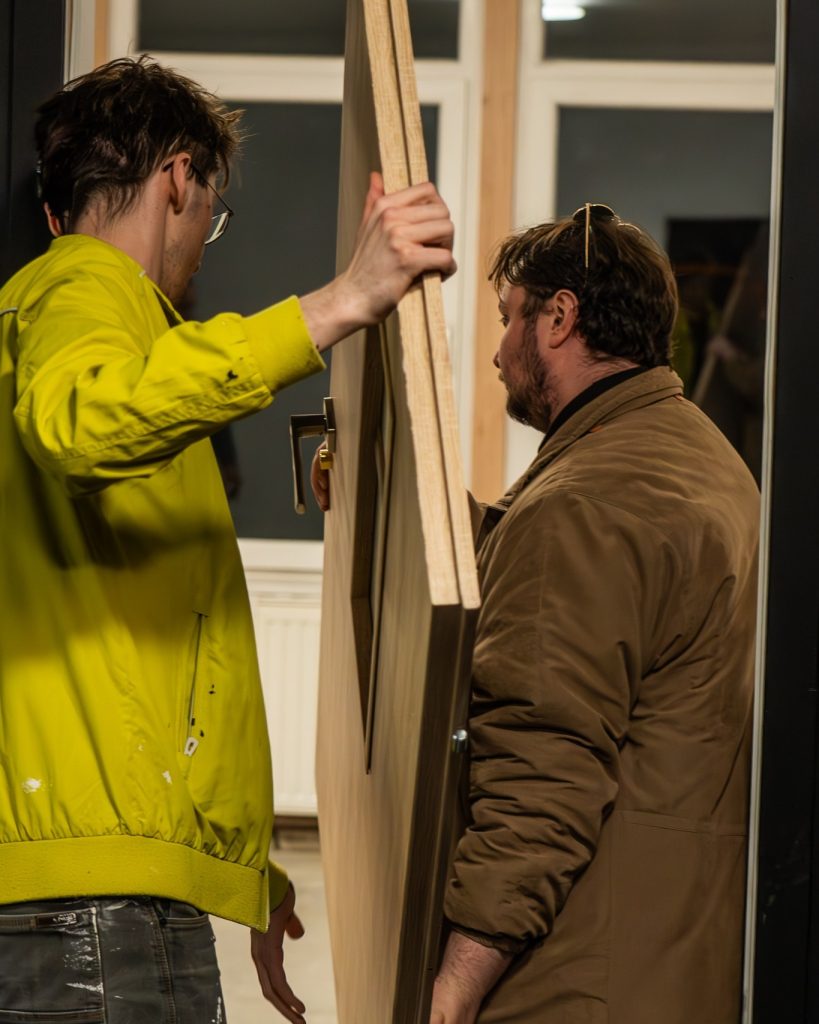 Dwóch mężczyzn wnosi duże drewniane drzwi do wnętrza pomieszczenia; jeden ubrany jest w żółtą kurtkę, drugi w brązowy płaszcz. Remont trwa.