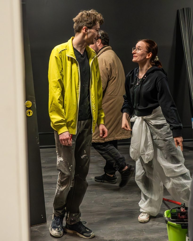 Trzy osoby w trakcie remontu; kobieta w czarnym stroju z białą folią ochronną rozmawia z mężczyzną w żółtej kurtce, w tle mężczyzna odchodzi z pomieszczenia.