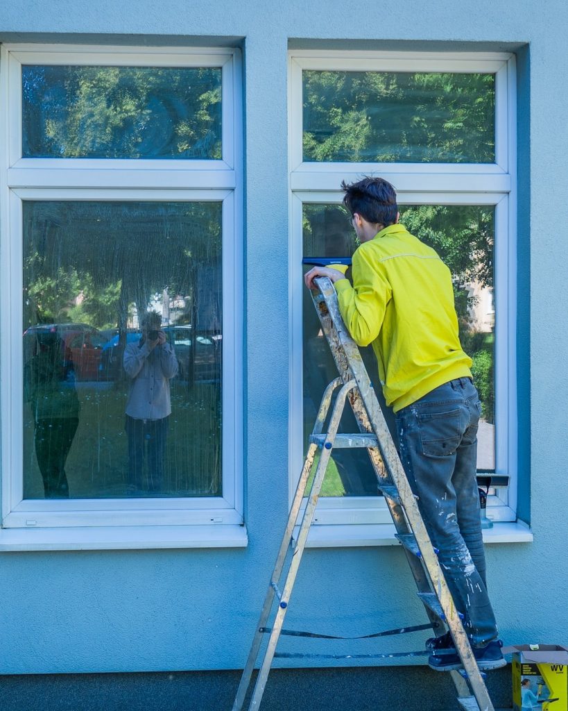 Mężczyzna w żółtej kurtce stoi na drabinie i czyści zewnętrzne okno budynku; w szybie odbija się osoba robiąca zdjęcie. Remont trwa.
