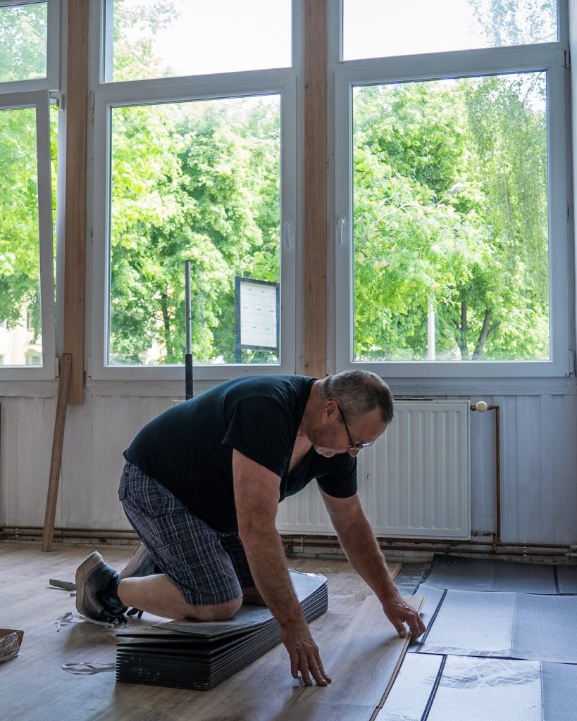 Starszy mężczyzna w okularach układa panele podłogowe w jasnym pomieszczeniu z dużymi oknami; klęczy na podłodze, precyzyjnie dopasowując elementy.