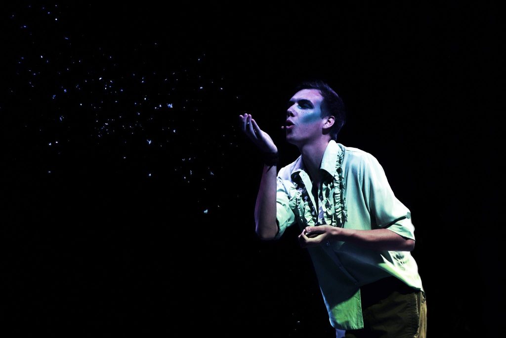 Mężczyzna na scenie w teatralnym makijażu, w jasnej koszuli z falbanami, dmucha w dłoń, rozsiewając drobinki w świetle reflektorów.