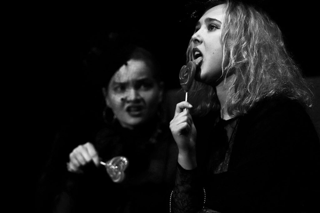 Dwie kobiety w teatralnym przedstawieniu, obie trzymają lizaki w kształcie serca; jedna z nich z grymasem niezadowolenia, druga zmysłowo liże lizaka.