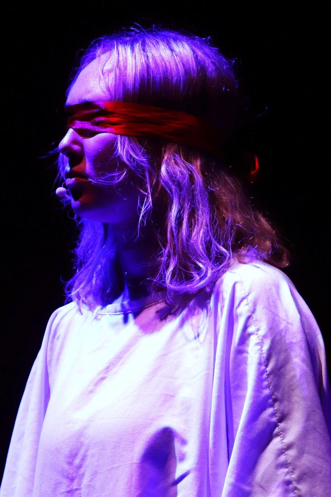 Młoda kobieta w białej koszuli z czerwoną opaską na oczach stoi na scenie w fioletowym świetle, z mikrofonem przy twarzy.