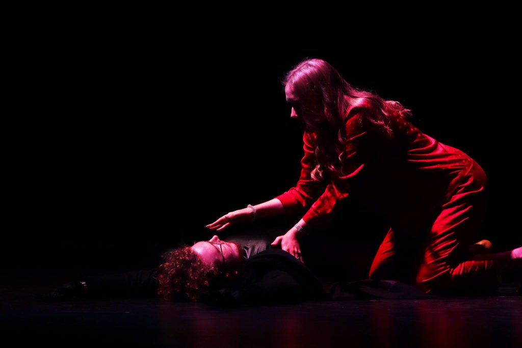 Kobieta w czerwonym kostiumie klęczy przy mężczyźnie leżącym na scenie; delikatnie dotyka jego klatki piersiowej i ramienia w dramatycznej scenie