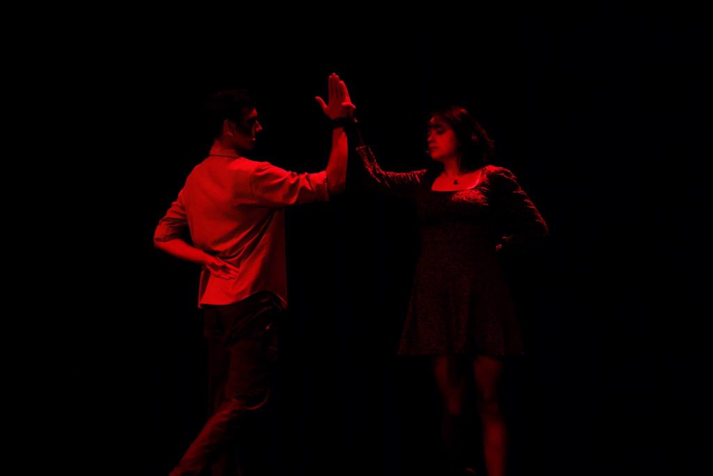 Mężczyzna i kobieta naprzeciw siebie na scenie, unoszą dłonie do wspólnego dotyku w geście napięcia emocjonalnego, oświetleni czerwonym światłem.