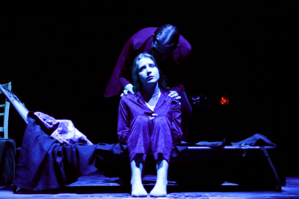 Kobieta w piżamie siedzi na łóżku teatralnym, patrząc w górę z melancholią, podczas gdy druga postać trzyma ją za ramiona od tyłu.