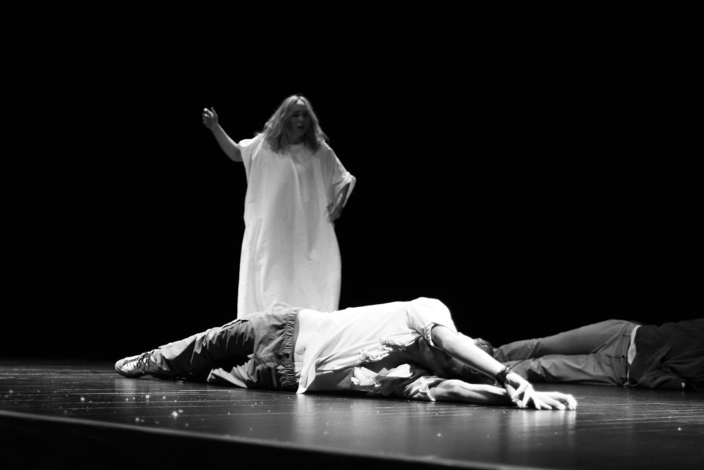Postać w białej sukni stoi na scenie, patrząc na dwie osoby leżące na podłodze w dramatycznej pozycji; scena utrzymana w czarno-białym tonie.