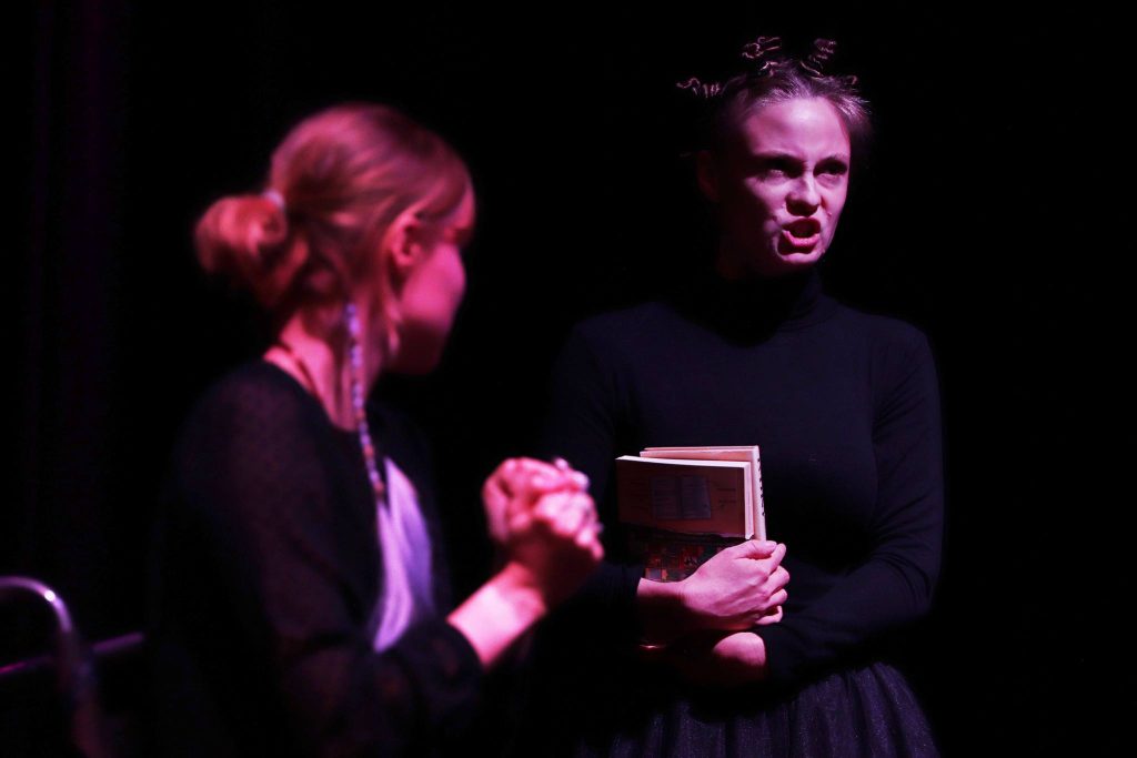 Dwie kobiety na scenie w intensywnym świetle – jedna z wyrazem gniewu trzyma książkę, druga stoi z złożonymi rękami, patrząc z niepokojem.