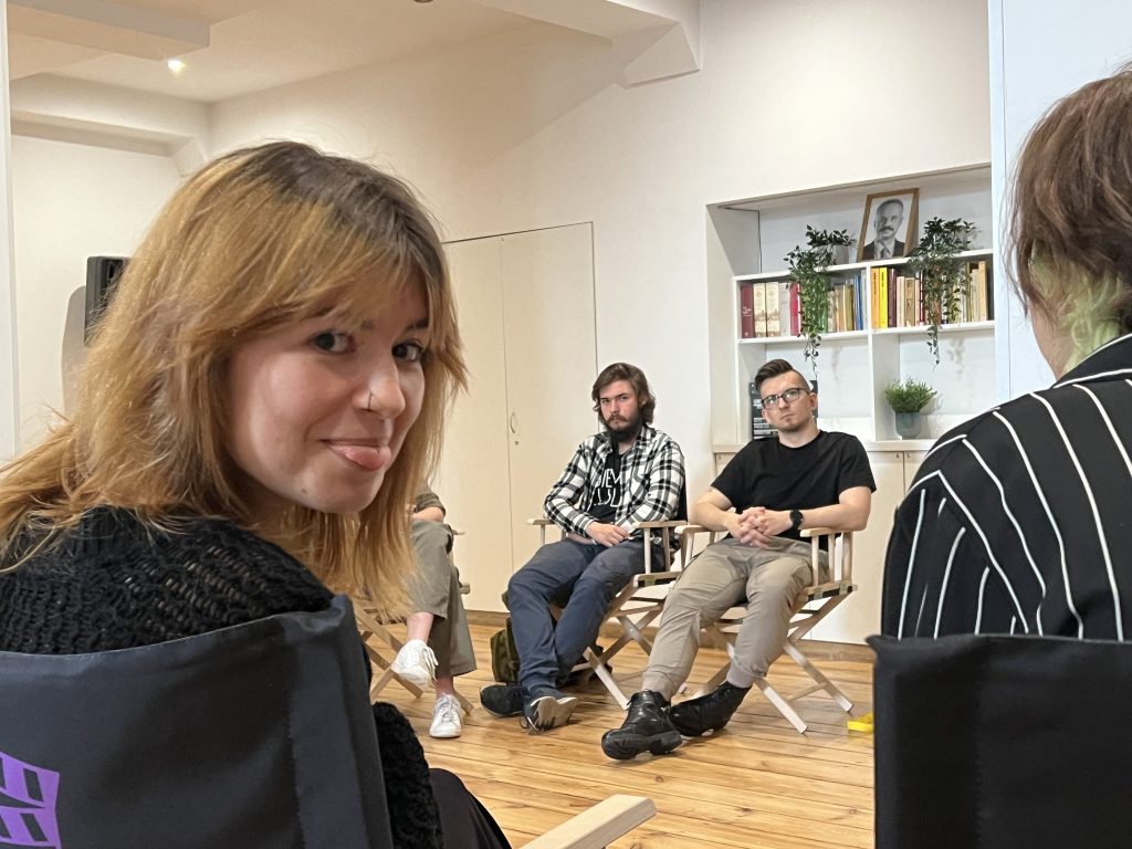 Młoda kobieta siedzi tyłem do aparatu i pokazuje język w stronę obiektywu, w tle widać trzech mężczyzn siedzących na krzesłach reżyserskich i słuchających rozmowy. Pomieszczenie ma jasne ściany i regał z książkami.