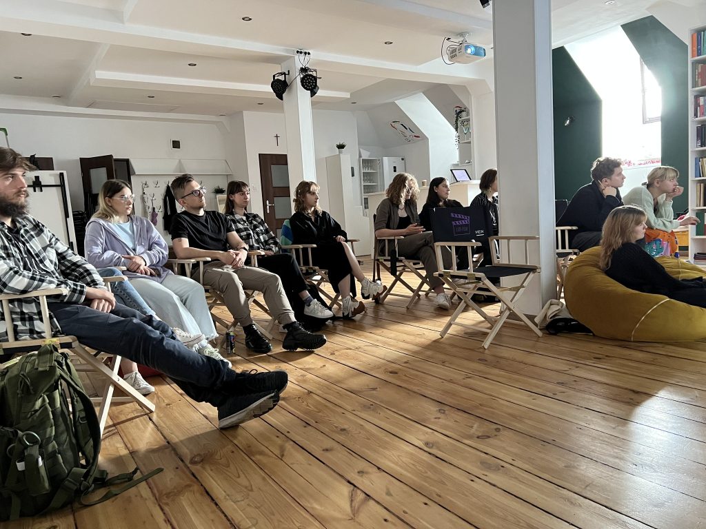 rupa uczestników warsztatów siedzi w półkolu na drewnianej podłodze przestrzeni warsztatowej. Niektórzy siedzą na krzesłach, inni na pufach. W tle regały z książkami i projektor sufitowy.