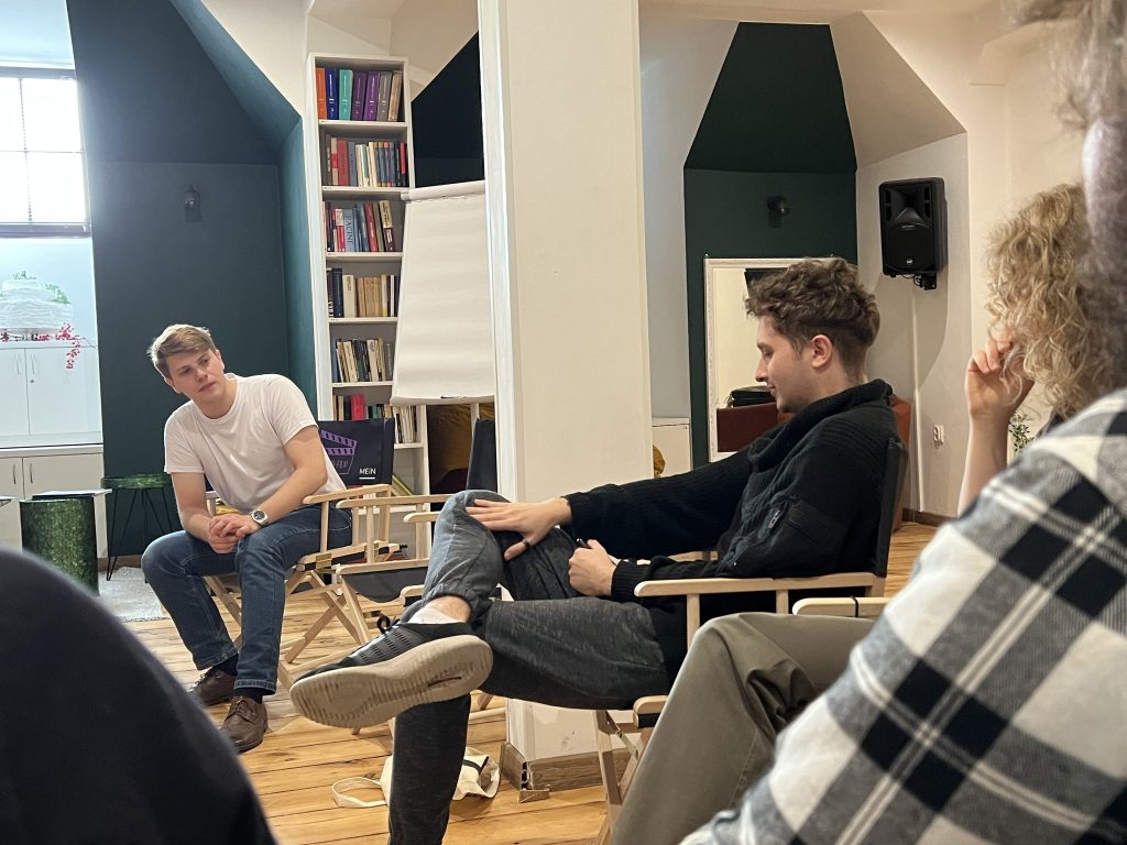 Dwóch młodych mężczyzn prowadzi rozmowę podczas zajęć. Jeden z nich siedzi bokiem z nogą założoną na nogę, drugi opiera się na krześle i uważnie słucha. W tle biblioteczka i duży arkusz papieru na stojaku.