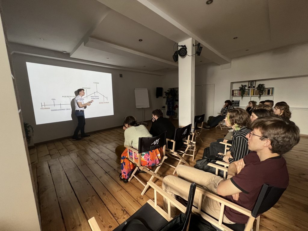 Mężczyzna w białej koszulce prowadzi prezentację, wskazując na diagram wyświetlany na ścianie. Uczestnicy siedzą w kilku rzędach na krzesłach reżyserskich, zwróceni w jego stronę.