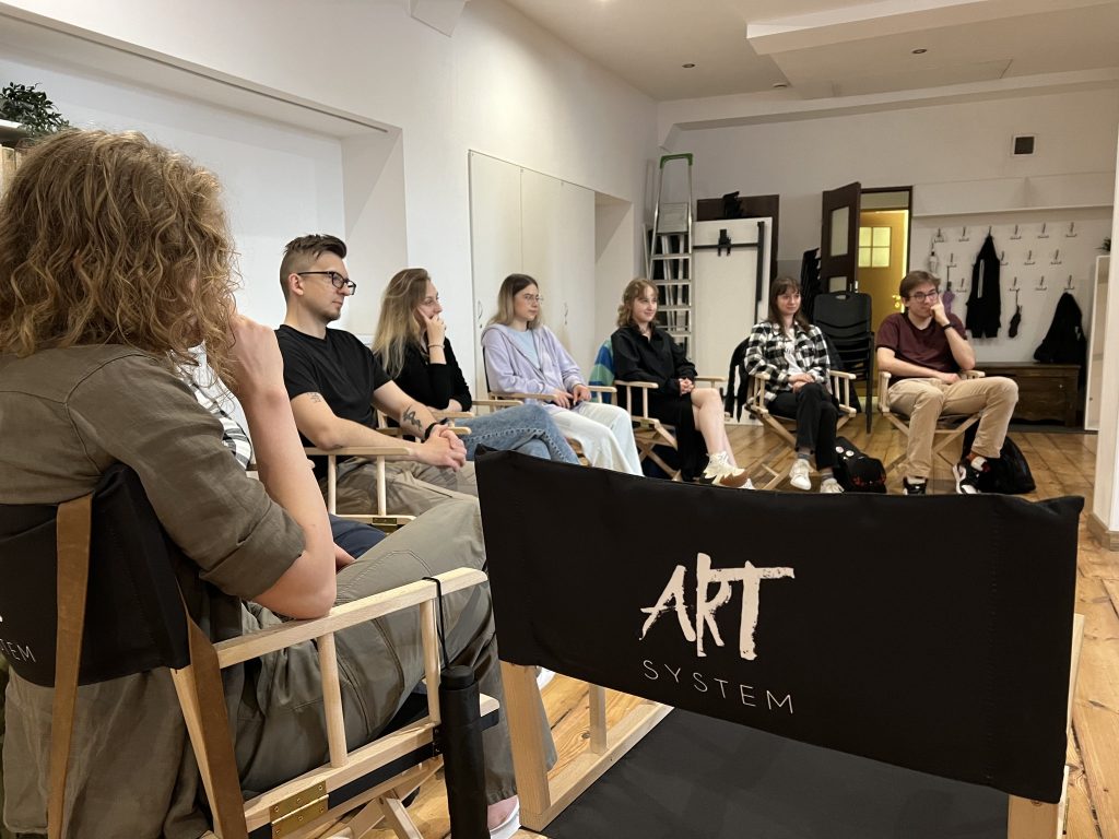 Grupa młodych osób siedzi w półkolu na warsztatach, skupiona na rozmowie. Na pierwszym planie widoczny jest nadruk 'ART SYSTEM' na oparciu krzesła. W tle widać drzwi, wieszaki i schowaną drabinę.