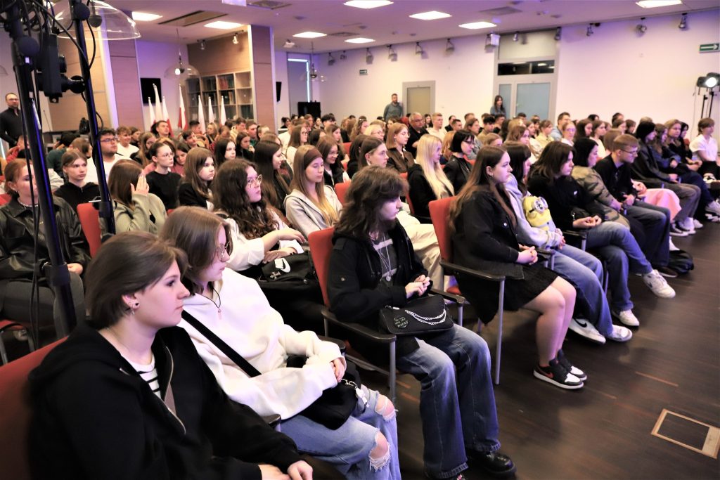 Publiczność złożona głównie z młodzieży siedząca na czerwonych krzesłach w sali konferencyjnej, uważnie śledząca wydarzenie.