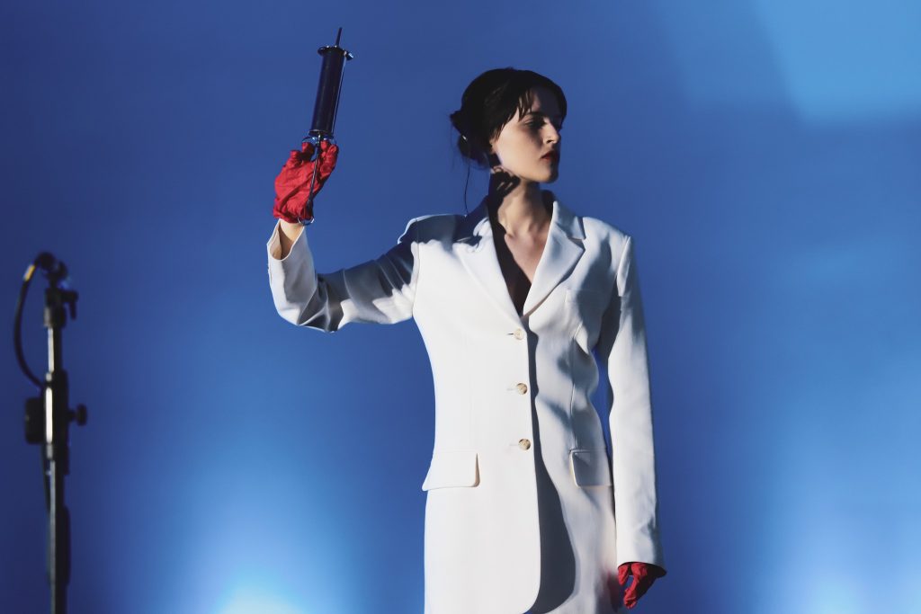 Kobieta w białym garniturze i czerwonych rękawiczkach trzymająca teatralnie dużą strzykawkę na tle niebieskiego światła.