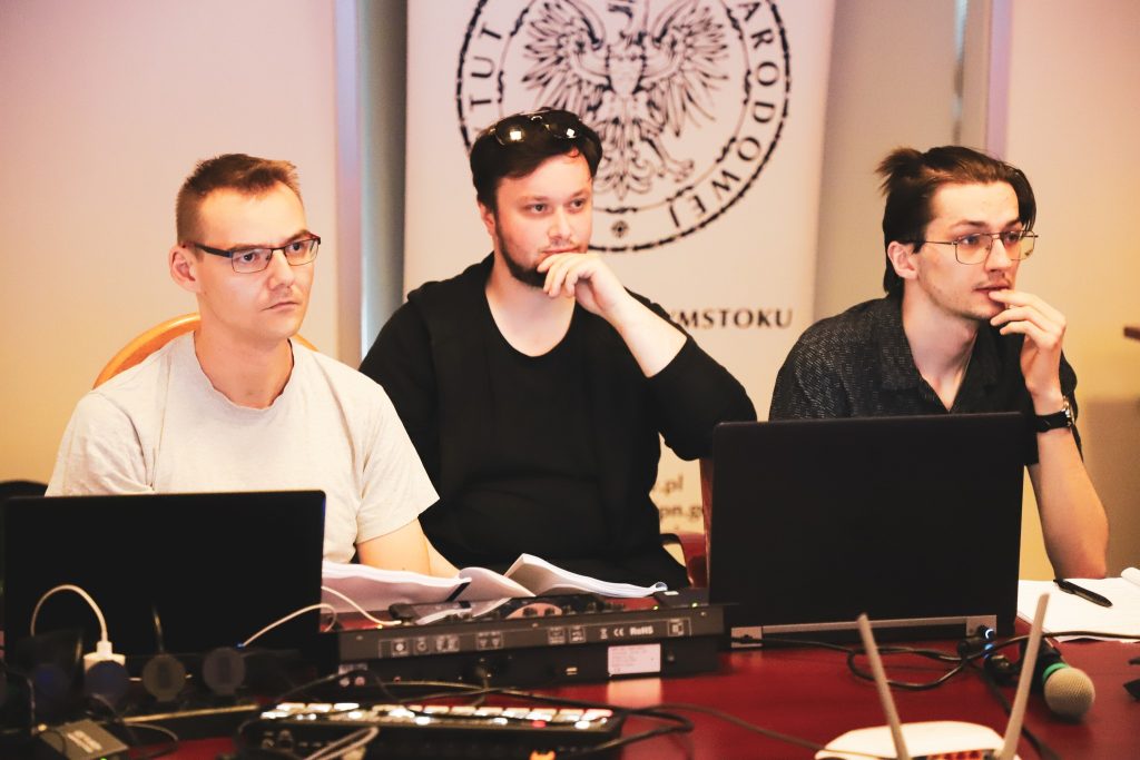 Trzech młodych mężczyzn skupionych przy stole z laptopami i dokumentami, obsługujących spektakl.