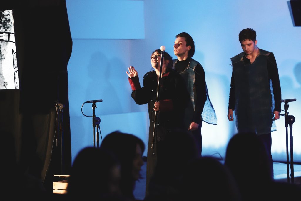 Trzy osoby na scenie – kobieta z pałką inscenizacyjną i dwaj młodzi mężczyźni w kostiumach z surowego materiału. Za nimi jasnoniebieskie tło. Na pierwszym planie widoczna jest publiczność.