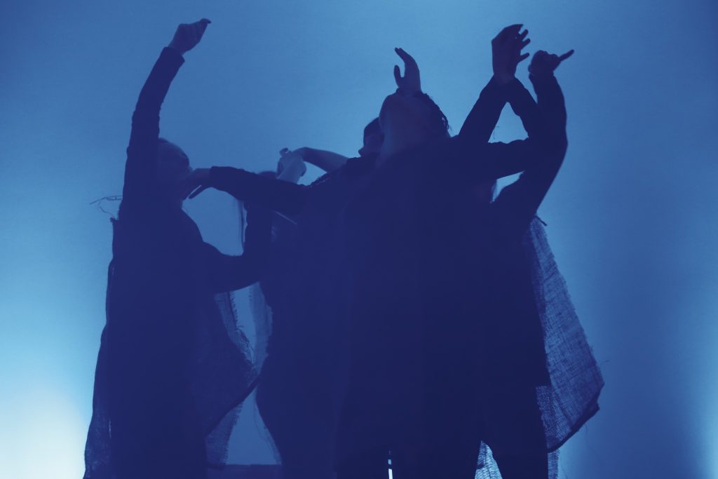 Grupa aktorów ubranych na czarno wykonuje ekspresyjny, synchroniczny ruch sceniczny z uniesionymi rękami. Tło wypełnia intensywne niebieskie światło, a sylwetki zlewają się z mroczną atmosferą.