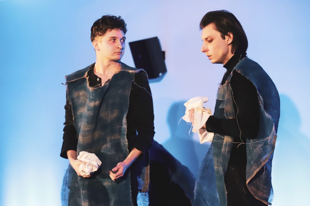 Dwóch młodych mężczyzn w teatralnych kostiumach przypominających worki z juty spogląda na siebie z powagą. Trzymają w dłoniach kawałki białego materiału, co sugeruje trudne emocjonalnie sceny.