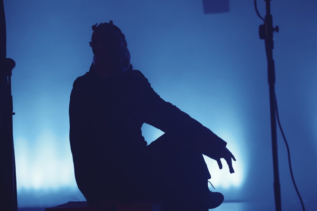 Sylwetka osoby w masce gazowej siedzącej w teatralnym półmroku na tle chłodnego, niebieskiego światła. Postać jest niemal całkowicie zacieniona, co buduje atmosferę grozy i tajemnicy.