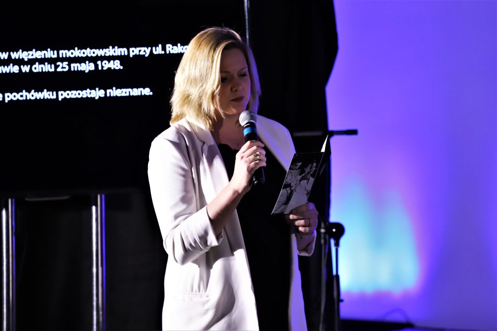 Kobieta w jasnej marynarce czyta tekst do mikrofonu na tle niebieskiego światła i wyświetlonego opisu historycznego.