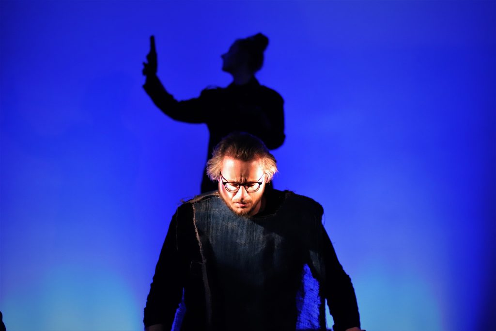 Mężczyzna w okularach i z poważnym wyrazem twarzy stoi na pierwszym planie w zniszczonym kostiumie. W tle widoczna kobieca postać w cieniu z uniesioną ręką, tworząc dramatyczny kontrast.