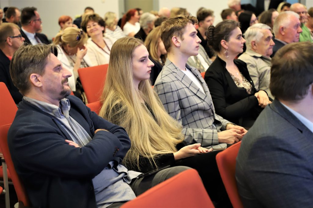 Grupa osób w różnym wieku siedząca w pierwszych rzędach sali konferencyjnej, skupiona na trwającym wydarzeniu.