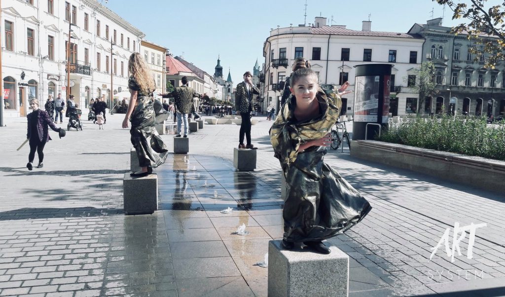 Młodzi artyści biorący udział w ulicznej akcji teatralnej ustawieni są na betonowych słupkach wzdłuż fontanny na deptaku w centrum miasta. Kobieta na pierwszym planie patrzy w stronę aparatu, pozując dynamicznie w połyskującym kostiumie. W tle widoczni są przechodnie i historyczna zabudowa miejska.