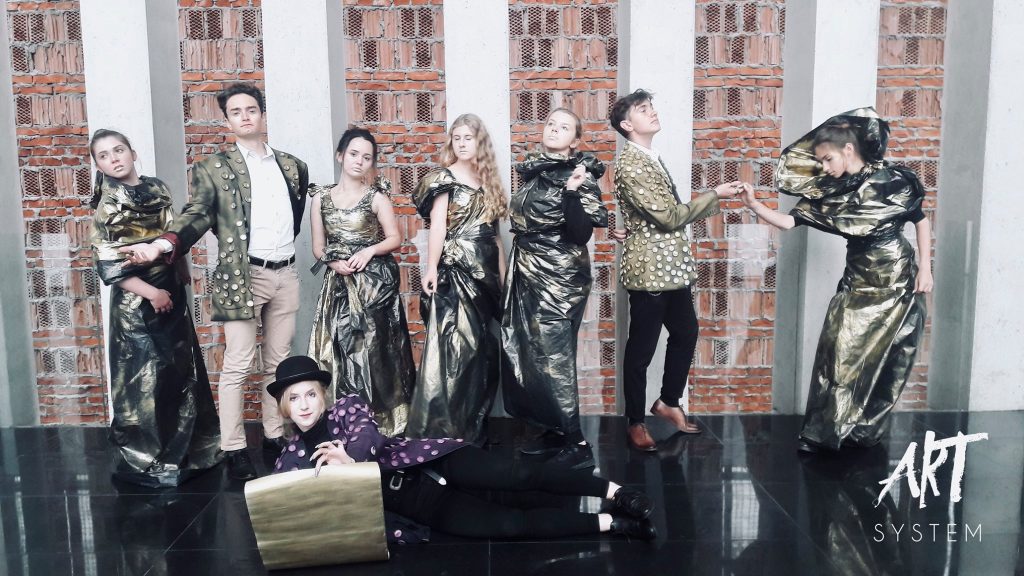 Artyści teatralni stoją w rzędzie na tle ceglanej ściany i białych filarów. Ubrani są w awangardowe stroje – suknie z metalicznego materiału i marynarki z dużymi guzikami. Na pierwszym planie, na podłodze, leży osoba w cylindrze i fioletowym płaszczu, spoglądając w stronę obiektywu.