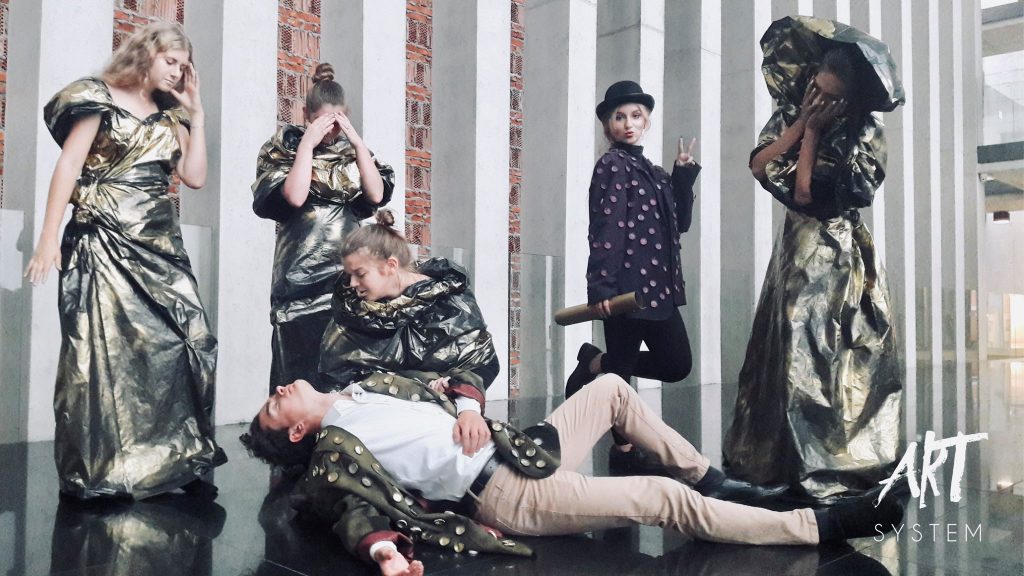 Scena dramatyczna – młody mężczyzna w zdobionej marynarce leży na podłodze jakby nieprzytomny, otoczony przez współperformerki w złotych kostiumach. Jedna z nich klęczy przy nim, a inna zakrywa oczy. Z boku aktorka w cylindrze wykonuje znak pokoju i uśmiecha się teatralnie.