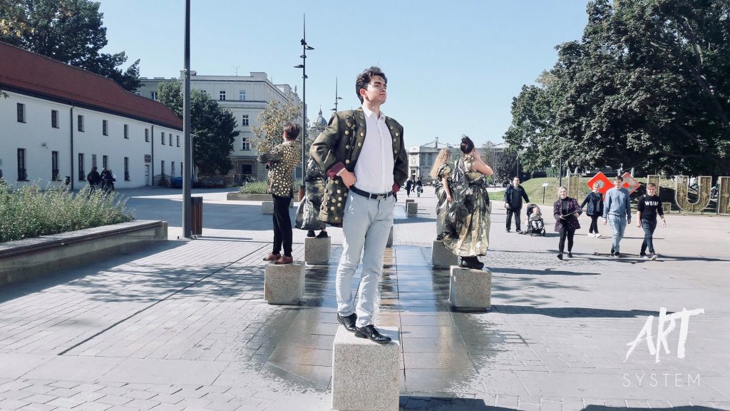 Performerzy stoją nieruchomo na niskich betonowych słupkach w przestrzeni miejskiej w słoneczny dzień. Na pierwszym planie mężczyzna w białej koszuli i marynarce z ozdobami patrzy dumnie w dal. Za nim widać przechodniów, budynki i drzewa.