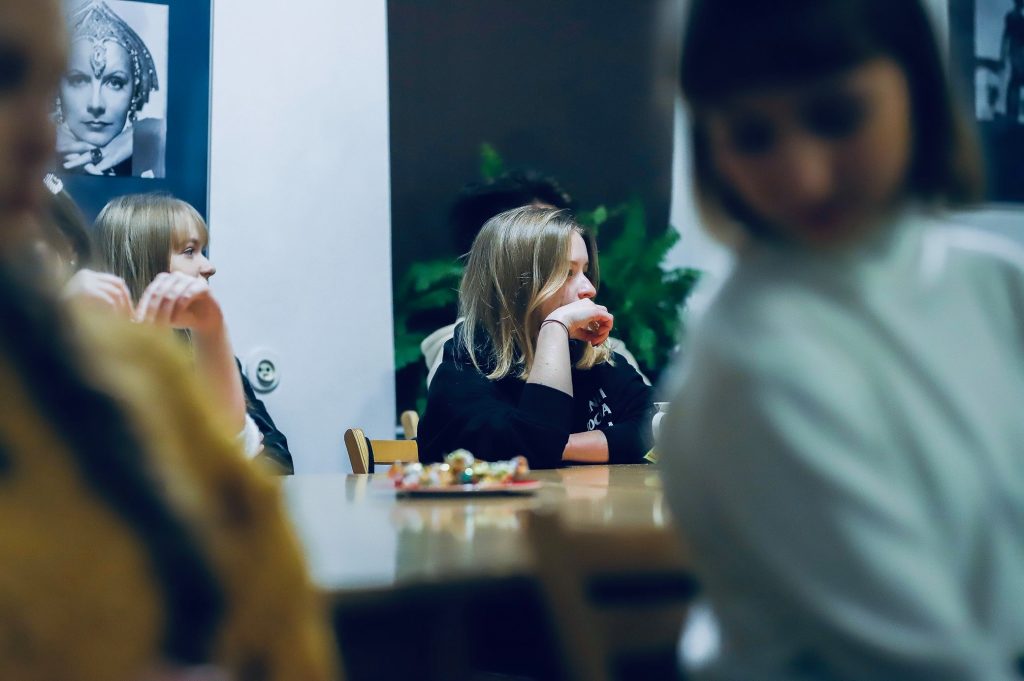 Kilka młodych osób siedzi przy stole, skupionych na rozmowie lub prezentacji; w tle widać czarno-białe zdjęcie aktorki w stylu retro.