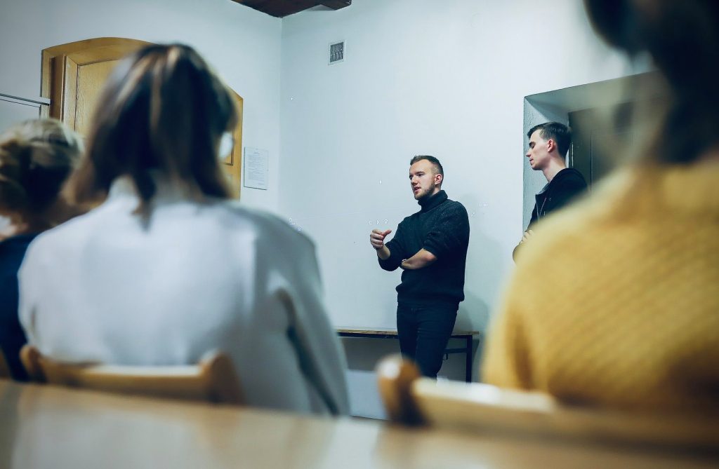 Dwóch młodych mężczyzn prowadzi spotkanie autorskie, stojąc przed grupą słuchaczy w kameralnym pomieszczeniu.