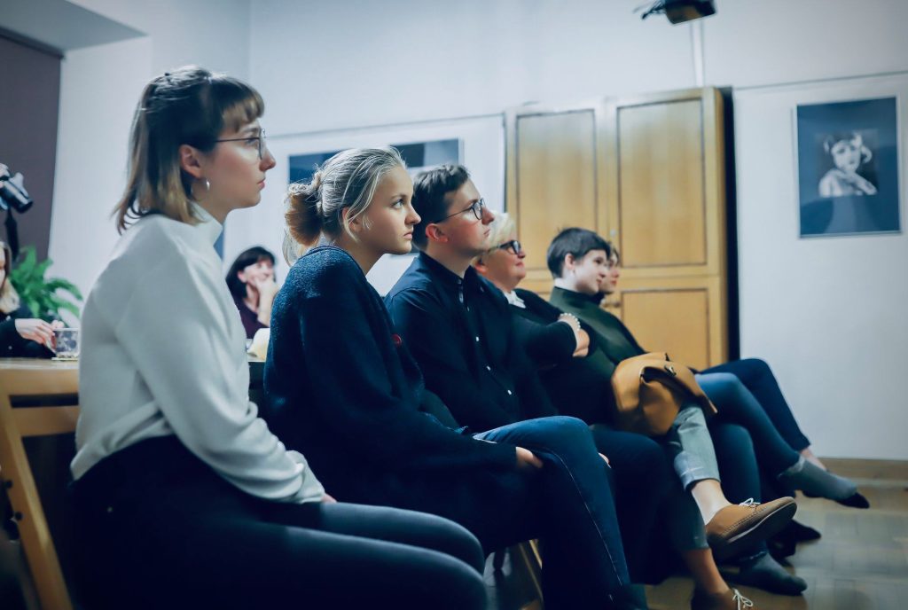 Uczestnicy pokazu filmu siedzą w skupieniu i słuchają prelekcji; sala ozdobiona jest czarno-białymi portretami aktorów.