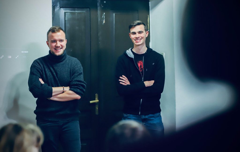 Dwóch prowadzących spotkanie autorskie stoi przy drzwiach z uśmiechami, z założonymi rękami, patrząc na publiczność.