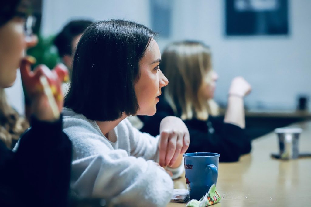 Młoda kobieta w białym swetrze siedzi przy stole z kubkiem i słucha uważnie rozmowy podczas pokazu filmu.