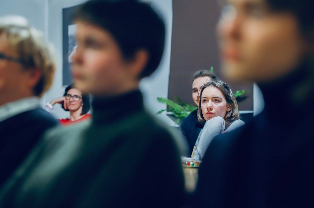 Kobieta w szarym swetrze patrzy w stronę prowadzącego z wyraźnym zaciekawieniem; wokół niej inni słuchacze.