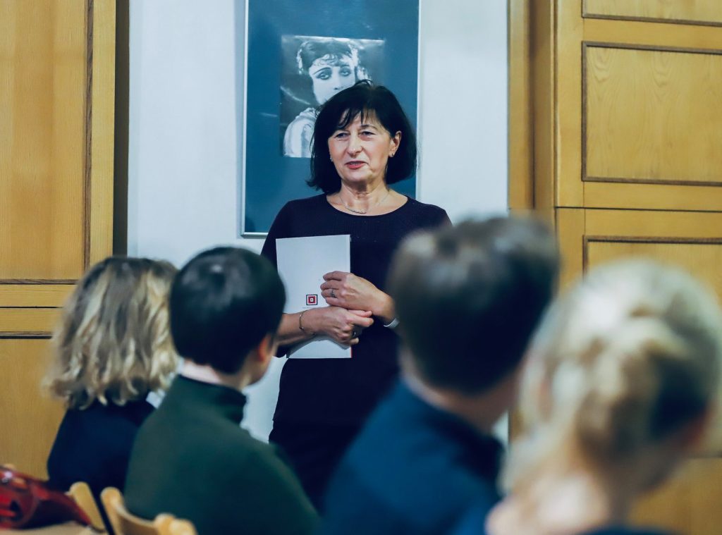 Kobieta z krótkimi ciemnymi włosami i teczką w ręku przemawia do zebranych uczestników w sali z portretami gwiazd kina.