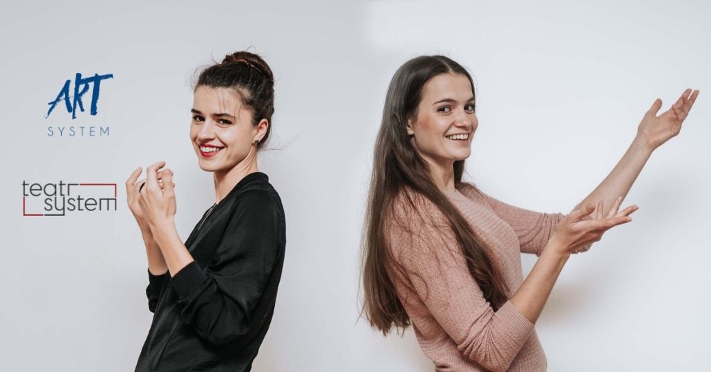 Dwie młode kobiety stoją tyłem do siebie na tle białej ściany, uśmiechnięte i wykonujące gesty rękami jakby coś prezentowały. Po lewej kobieta w czarnej bluzie z upiętymi włosami, po prawej kobieta w różowym swetrze z długimi rozpuszczonymi włosami. Na zdjęciu widoczne są także logotypy: niebieski „ART System” oraz czarno-czerwony „Teatr System”. Zdjęcie ma jasny, przyjazny charakter.